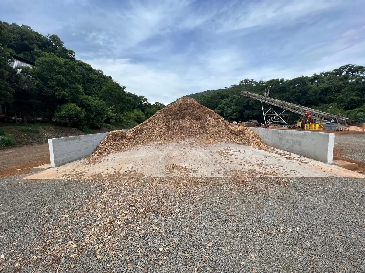 Baia de concreto armado concluída, com laje e muros, em uso com estoque de cavaco de madeira junto à correia transportadora.