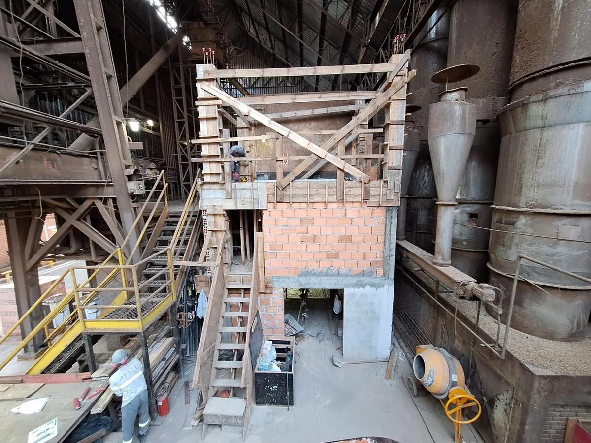 Pilares e vigas de concreto em fôrmas de madeira sobre alvenaria de vedação, entre ciclones e silos de uma área industrial, com profissional da Ápice em primeiro plano.