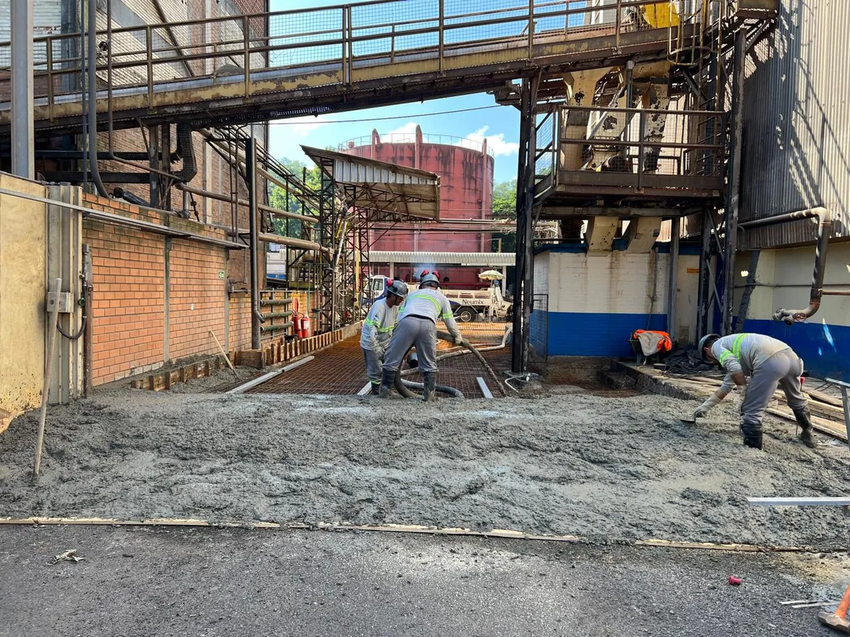 Equipe de capacete e uniforme refletivo lança concreto bombeado sobre a tela de aço do pavimento de uma via interna de fábrica, sob passarela metálica.