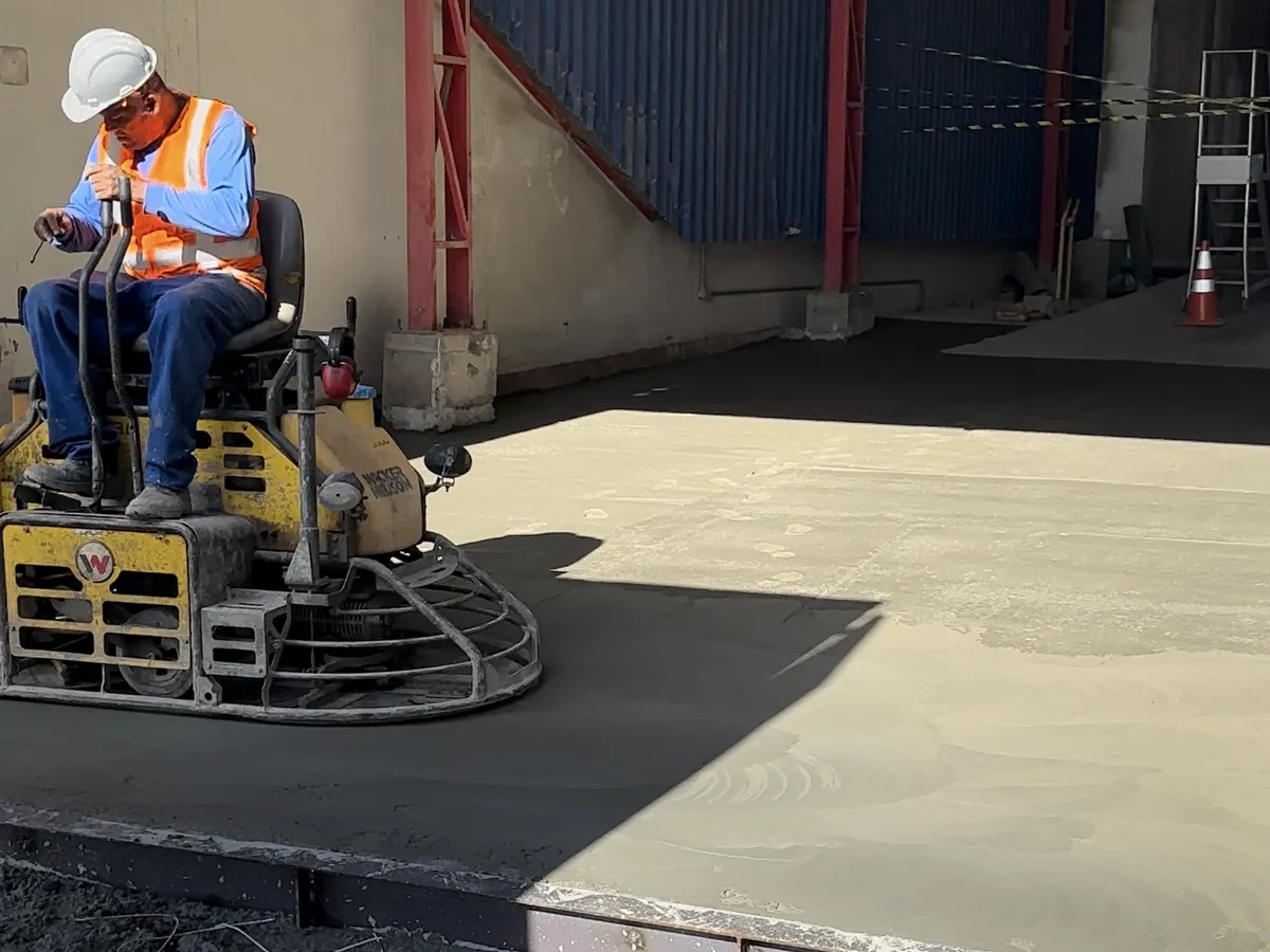 Operador de capacete e colete conduz acabadora mecânica dupla sobre o piso de concreto recém-lançado, na entrada de um galpão industrial.